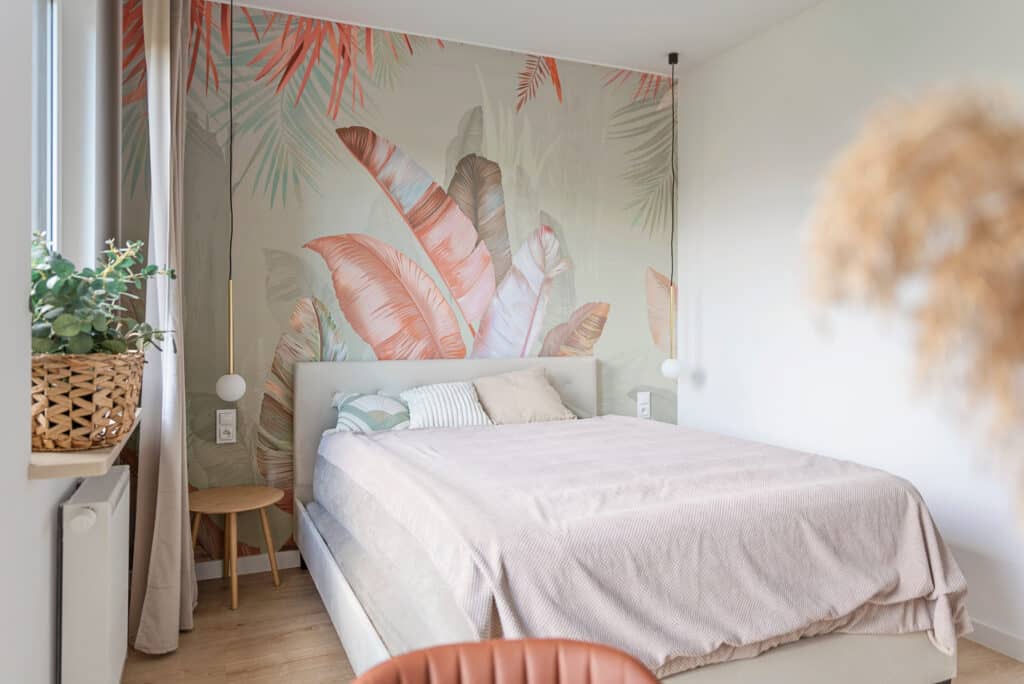 Chambre design moderne: lit confortable, déco tropicale feuilles. Chambre moderne avec lit double, tête de lit beige et couette rose. Papier peint tropical feuilles terracotta et vertes, plantes et luminaires.